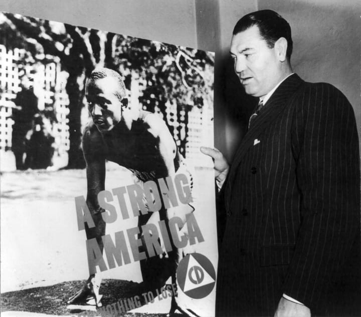 Portrait or photograph of Jesse Owens: American boxer Jack Dempsey posed with Civil Defense poster "A Strong America" showing Jesse Owens on starting blocks.