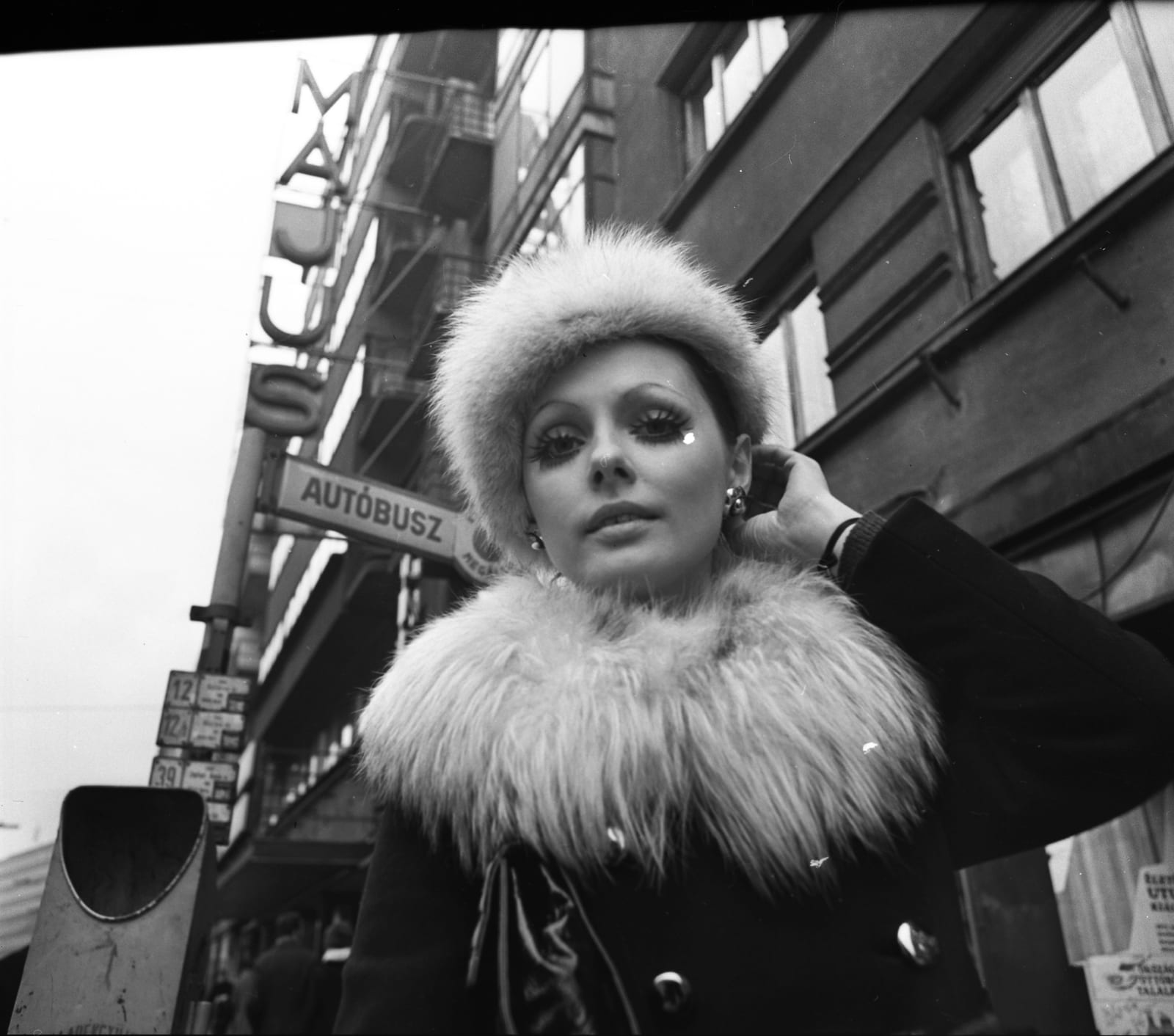 Portrait or photograph of Mozi: Szilágyi Mari, Margit körút (Mártírok útja), buszmegállóban, háttérben az 55. számú épületben a Május 1. mozi (ma Átrium Film-Sz